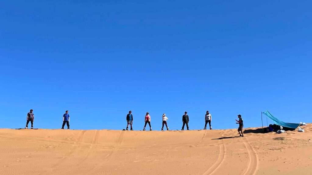 Grupo de 8 turistas practicando el sandboard, momento antes de deslizarse por la duna